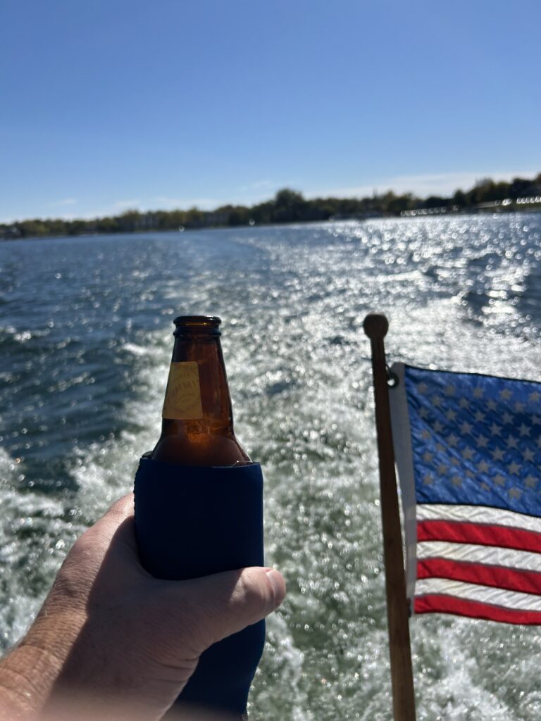 Person holding a cold beer in a navy koozie on a boat on Lake Minnetonka, with an American flag and sparkling wake in the background
